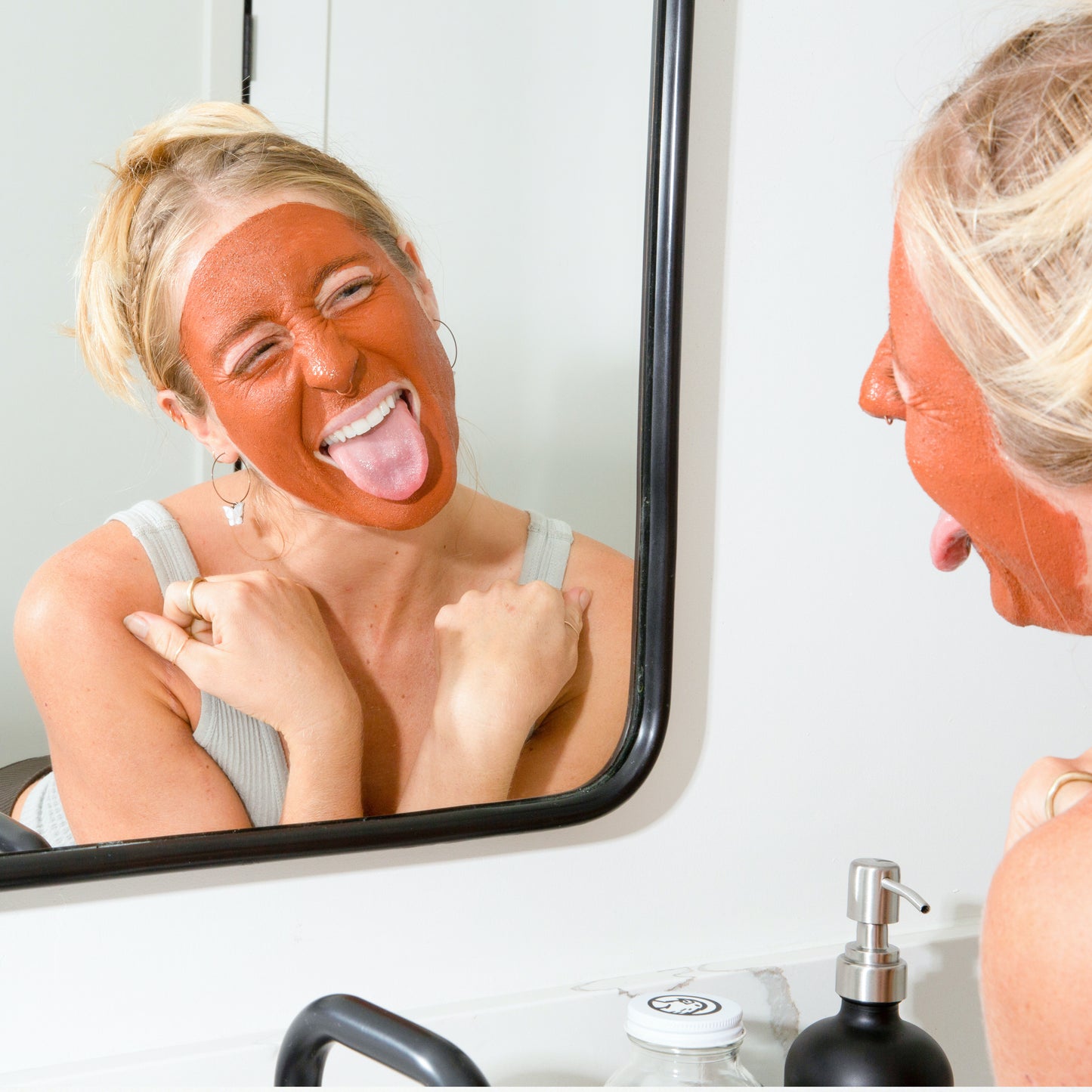 Woman making a silly face while using canary clay + rose face mask in bathroom
