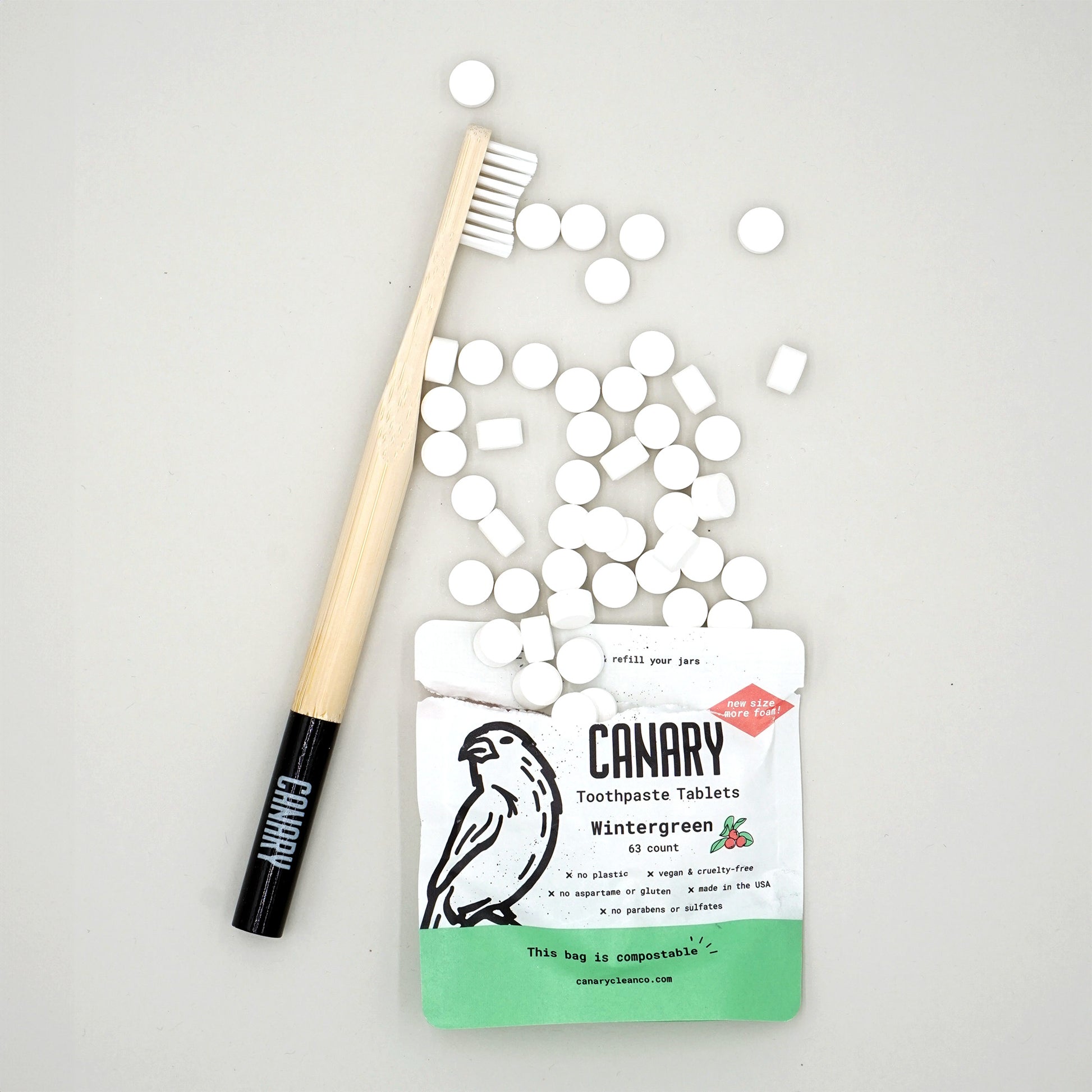 Canary Wintergreen Toothpaste Tablets, sample size 63 count compostable pouch, front view of the pouch with tablets spilling out  and with Canary toothbrush